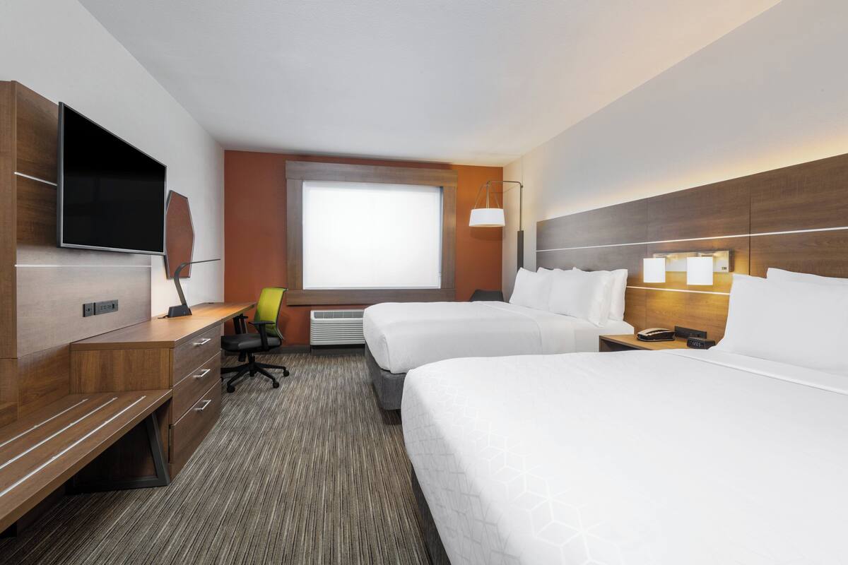 A hotel room with two beds, a desk with a chair, a flat-screen TV, and a large window.
