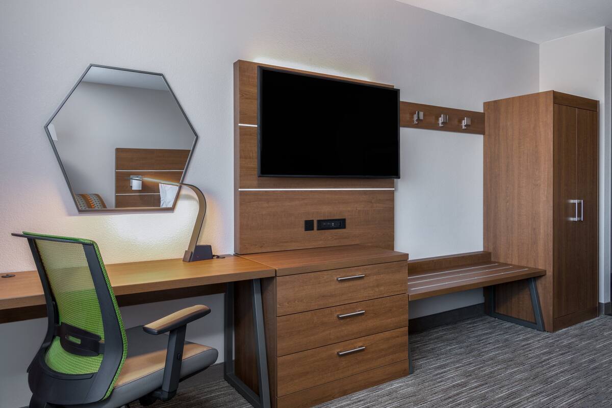 A modern hotel room with a wooden desk, a chair, a TV mounted on the wall, a mirror, and a wardrobe.