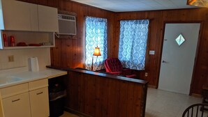 A compact kitchen with wooden walls, a white countertop, a red chair, and a round wooden table with a clock. - Stand alone cabin,  sanitized, full kitchen. (Cadiz)