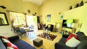 A spacious living room with a sofa set, a coffee table, and a TV. There is a dining area with chairs and a table, and a kitchen area with a stove and cabinets.