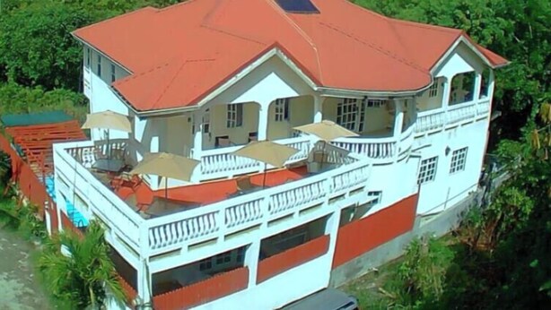 A large, two-story building with a red roof and white walls, surrounded by lush greenery and mountains in the background.