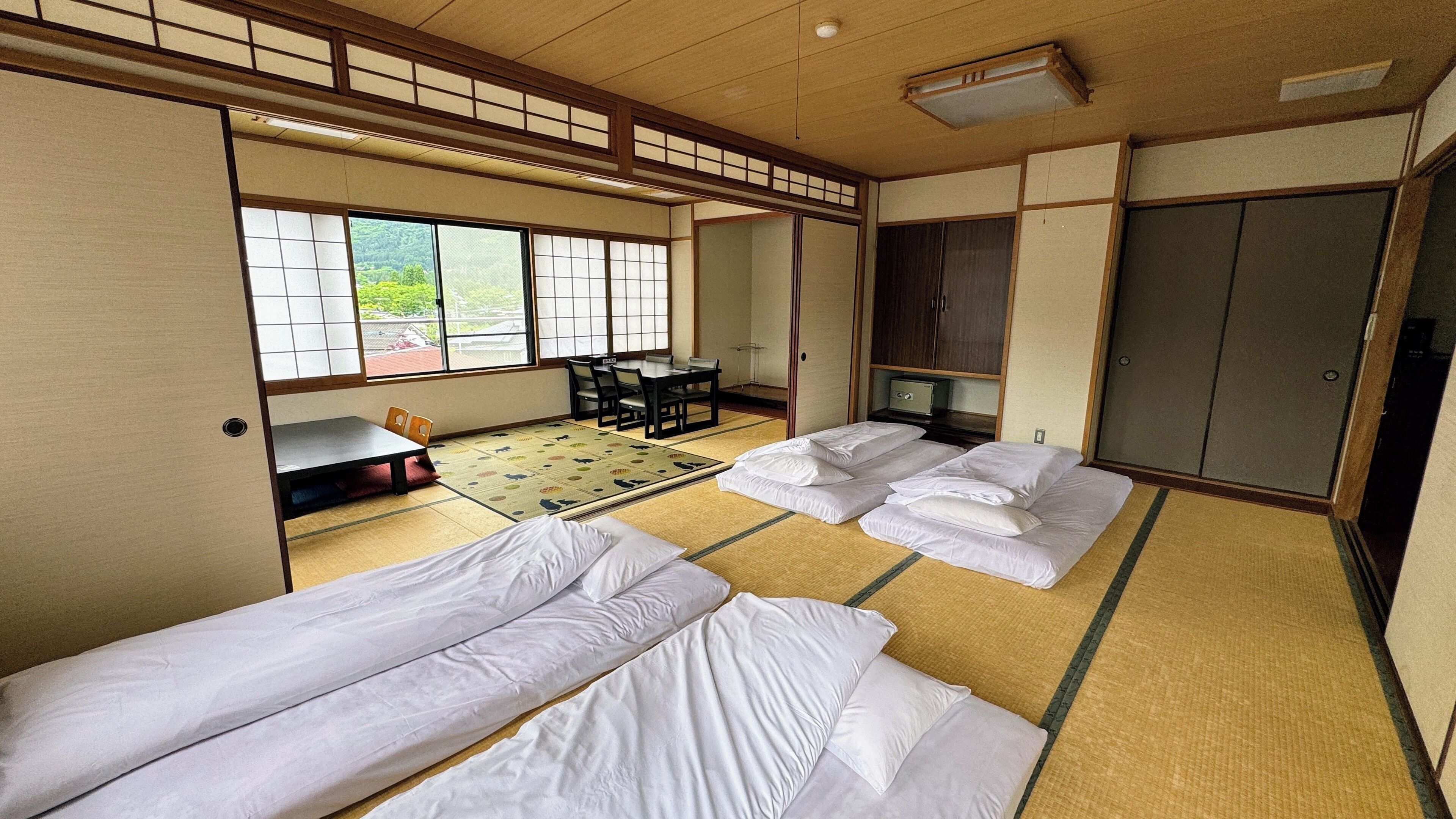 A traditional Japanese room with tatami flooring, two beds, a table with chairs, and a view of greenery through large windows.