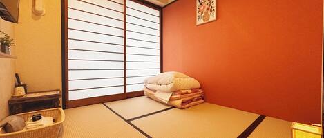 A traditional Japanese room with tatami flooring, a futon bed, a shoji screen, and a small shelf with personal items.