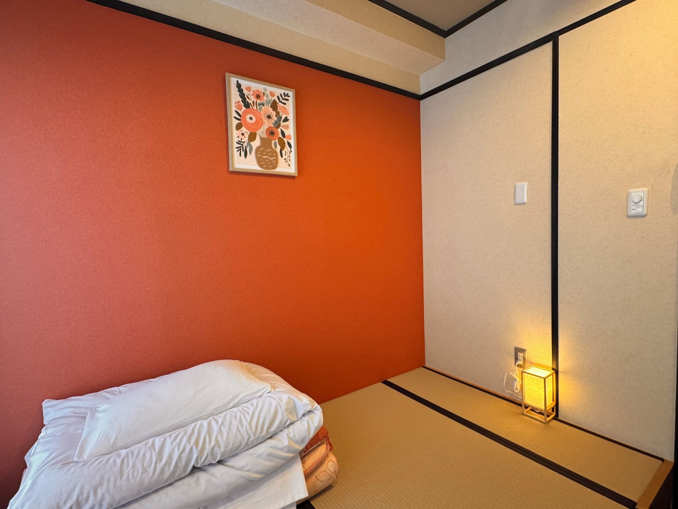 A room with an orange wall, a bed with white bedding, a tatami mat floor, a wall-mounted light, and a floral artwork.