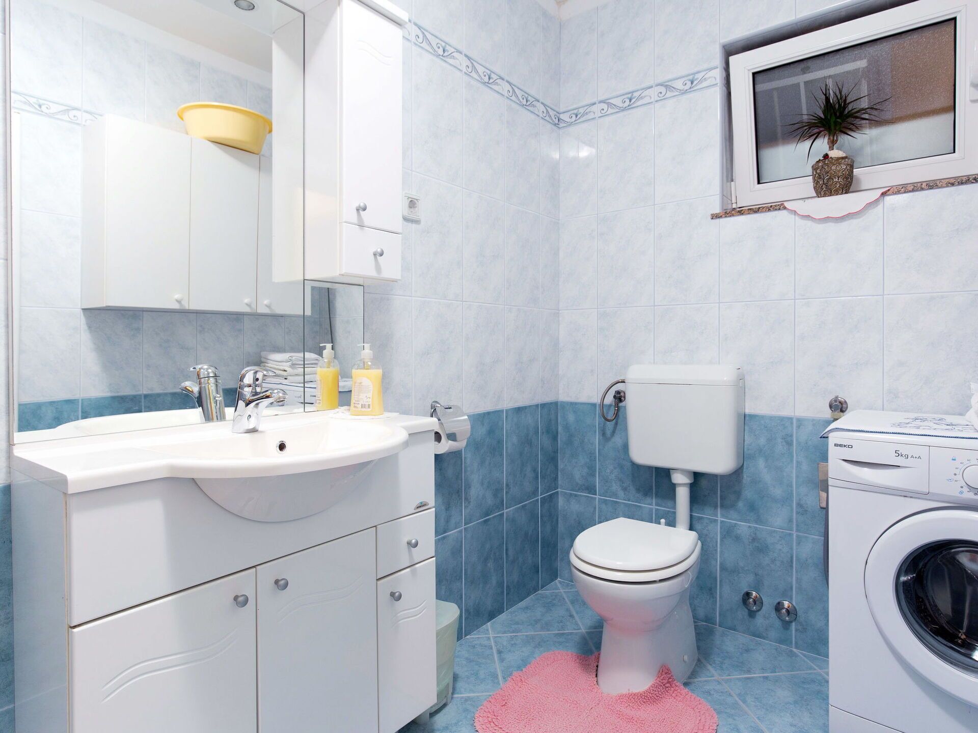 A modern bathroom with a white sink, mirror, and toilet, a washing machine, and a blue-tiled floor.
