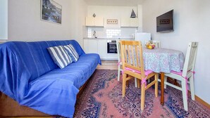 A compact living space with a blue sofa, a dining table with chairs, a kitchen area, and a patterned rug.