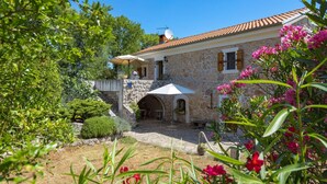 Un bâtiment en pierre avec un toit en tuiles, un patio couvert avec des sièges extérieurs et un mur en pierre surmonté d’une petite arche.