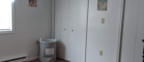 Quarto com janela, radiador, cesto de roupa suja e duas portas.