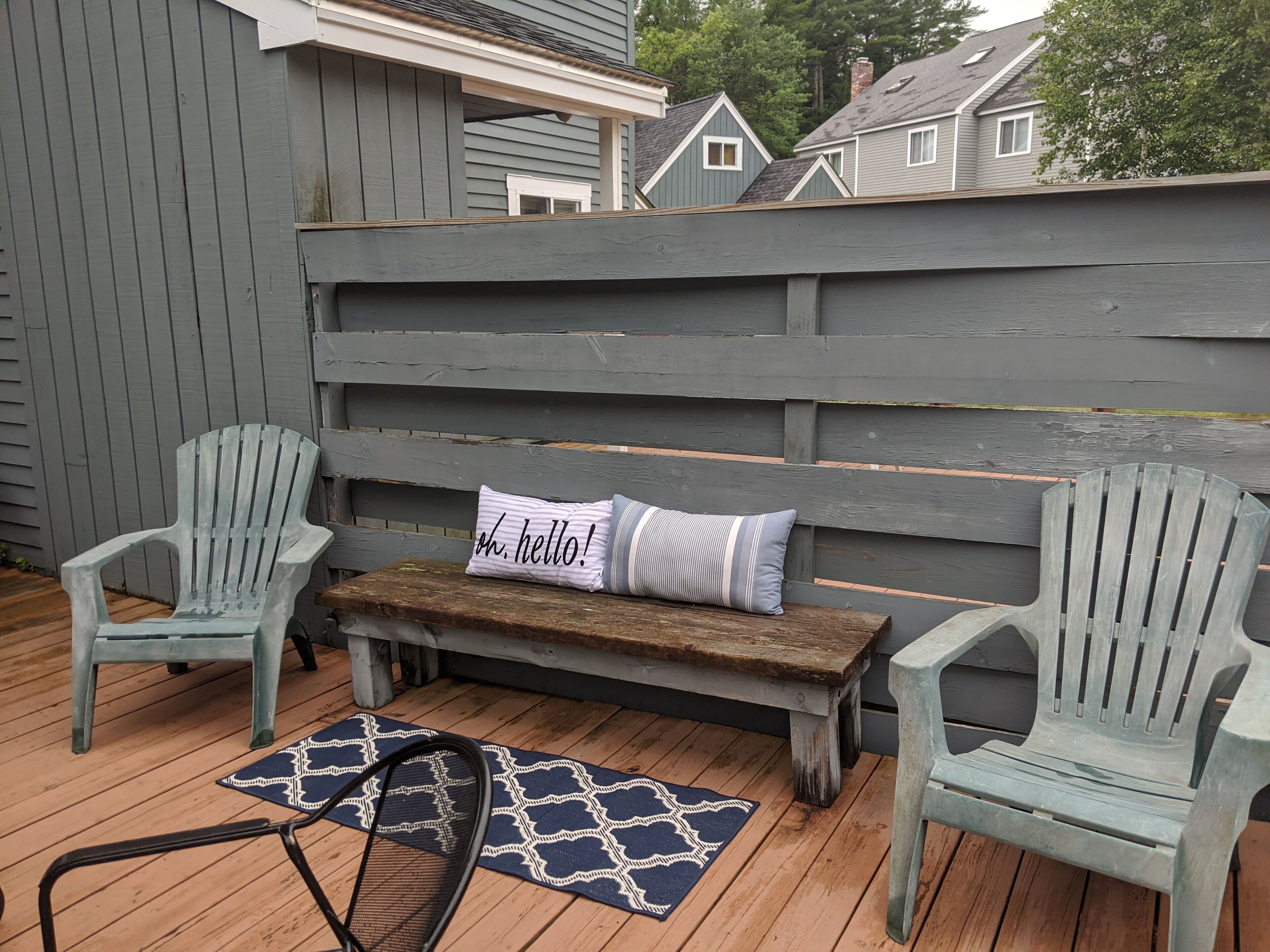A wooden deck with two Adirondack chairs, a bench, and a patterned rug.