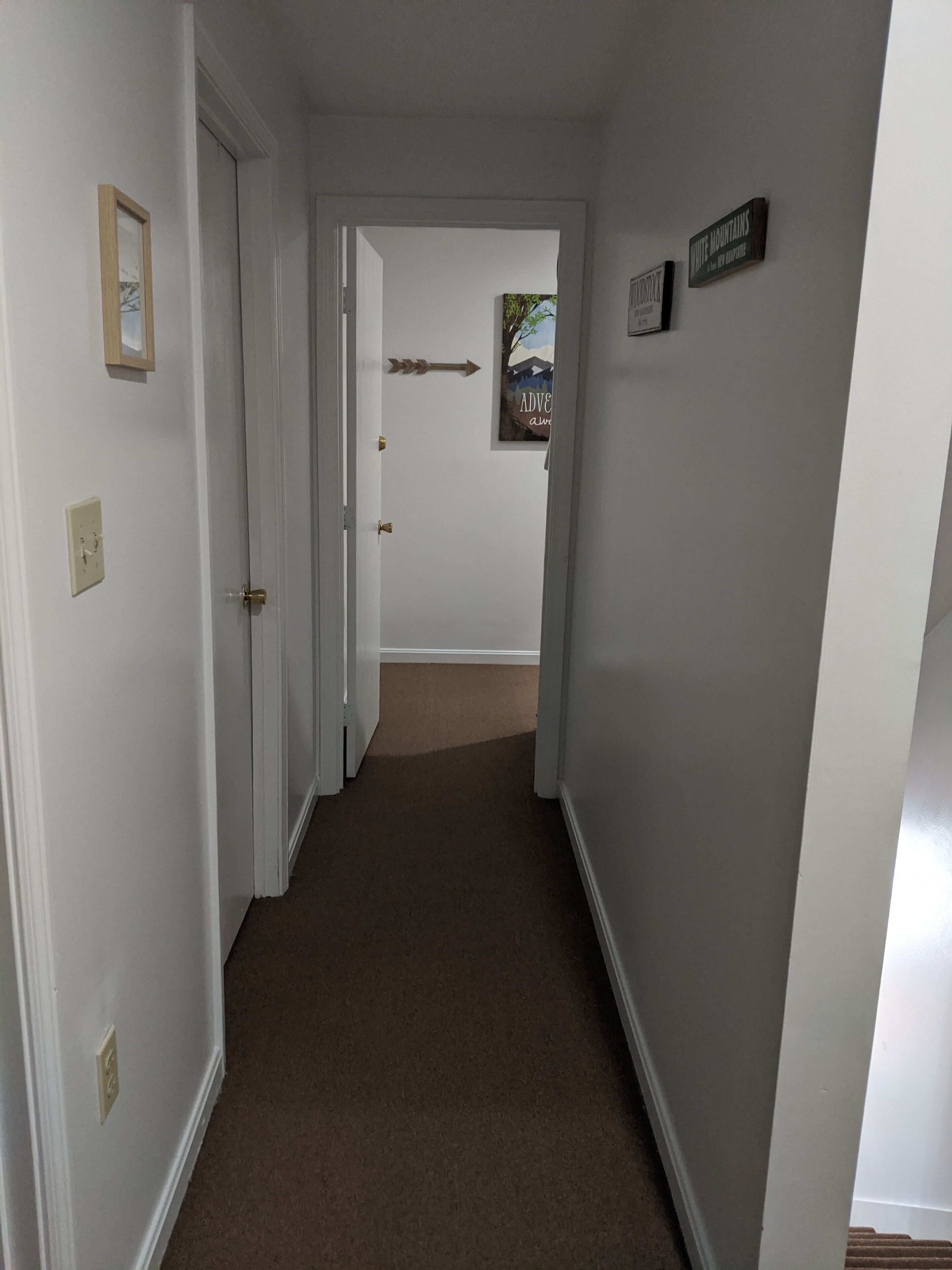 A narrow hallway with white walls and a brown carpet. There are doors on the left, a sign on the right, and a framed picture on the wall.