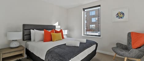 Un dormitorio moderno con una cama grande, una mesita de noche con lámpara, una silla y una ventana con vistas a edificios.