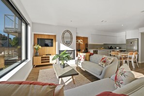 A modern living room with a grey sofa, a coffee table, and a TV. There is a kitchen area with a dining table and chairs. - Residence du Parc by Staysouth (Arthur's Point)