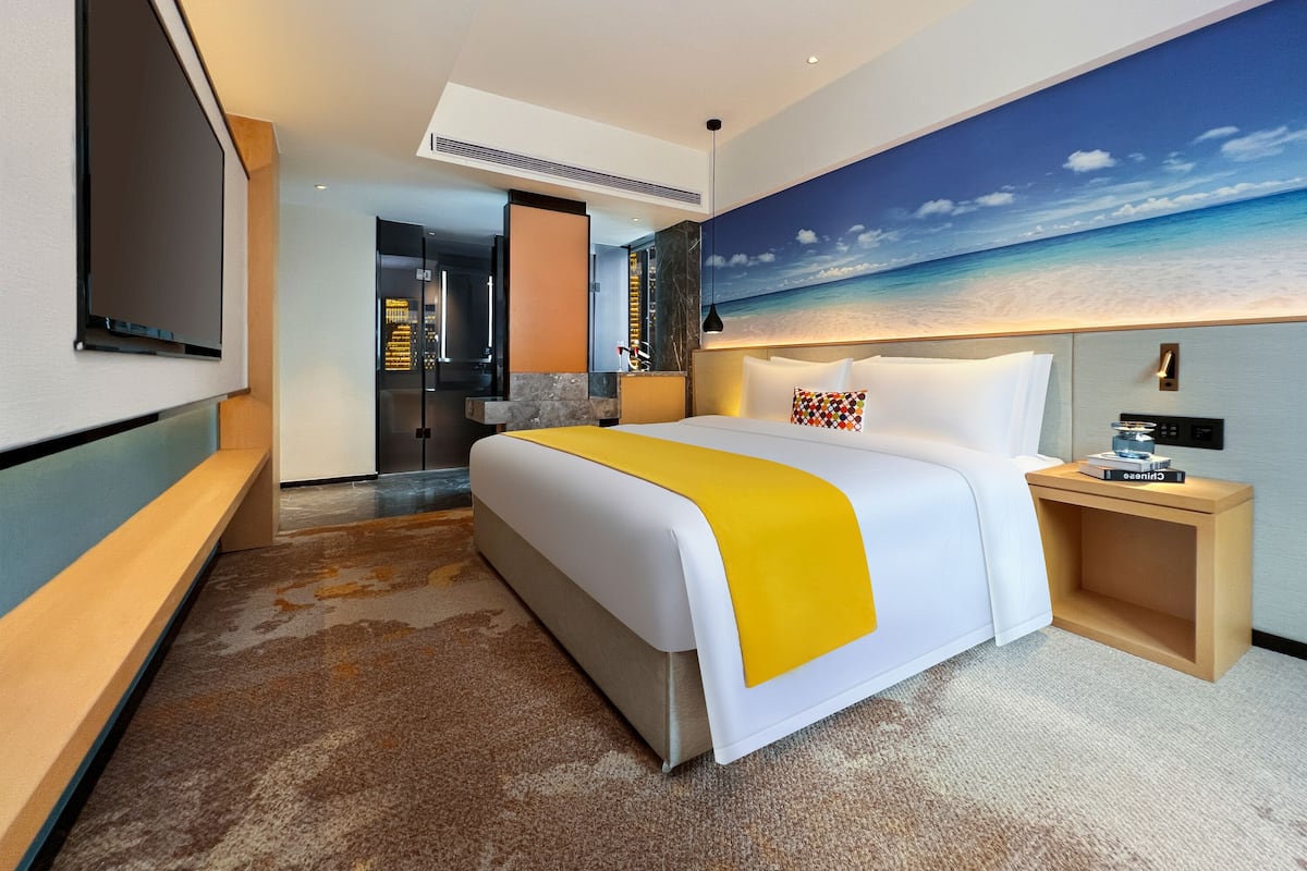 Ein modernes Hotelzimmer mit einem großen Bett, einem Fernseher, einem Strandbild und einem kleinen Holftisch.