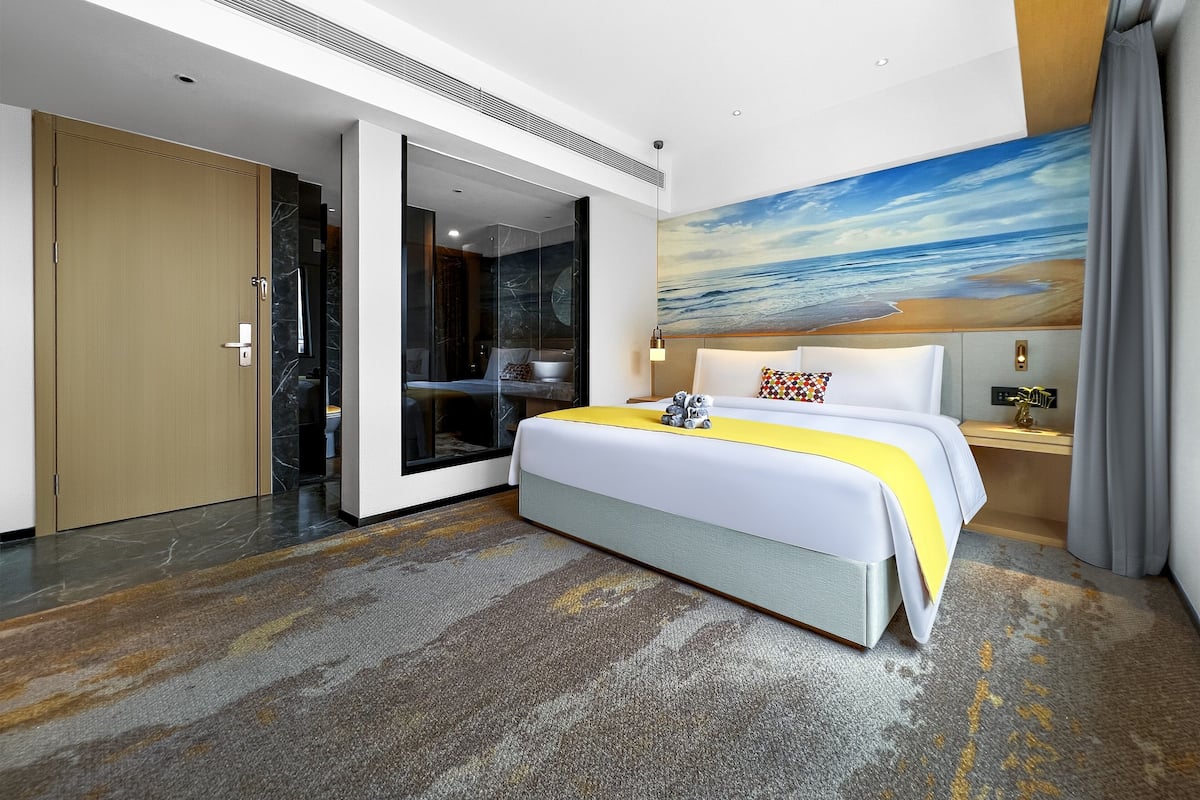 Ein Hotelzimmer mit einem großen Bett, einem Strandbild, einer Tür und einem Nachttisch mit einer Lampe.