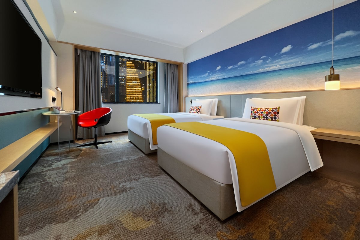 Ein Hotelzimmer mit zwei Betten, einem Schreibtisch, einem Stuhl, einem Fernseher und einem Wandbild mit Strandblick.