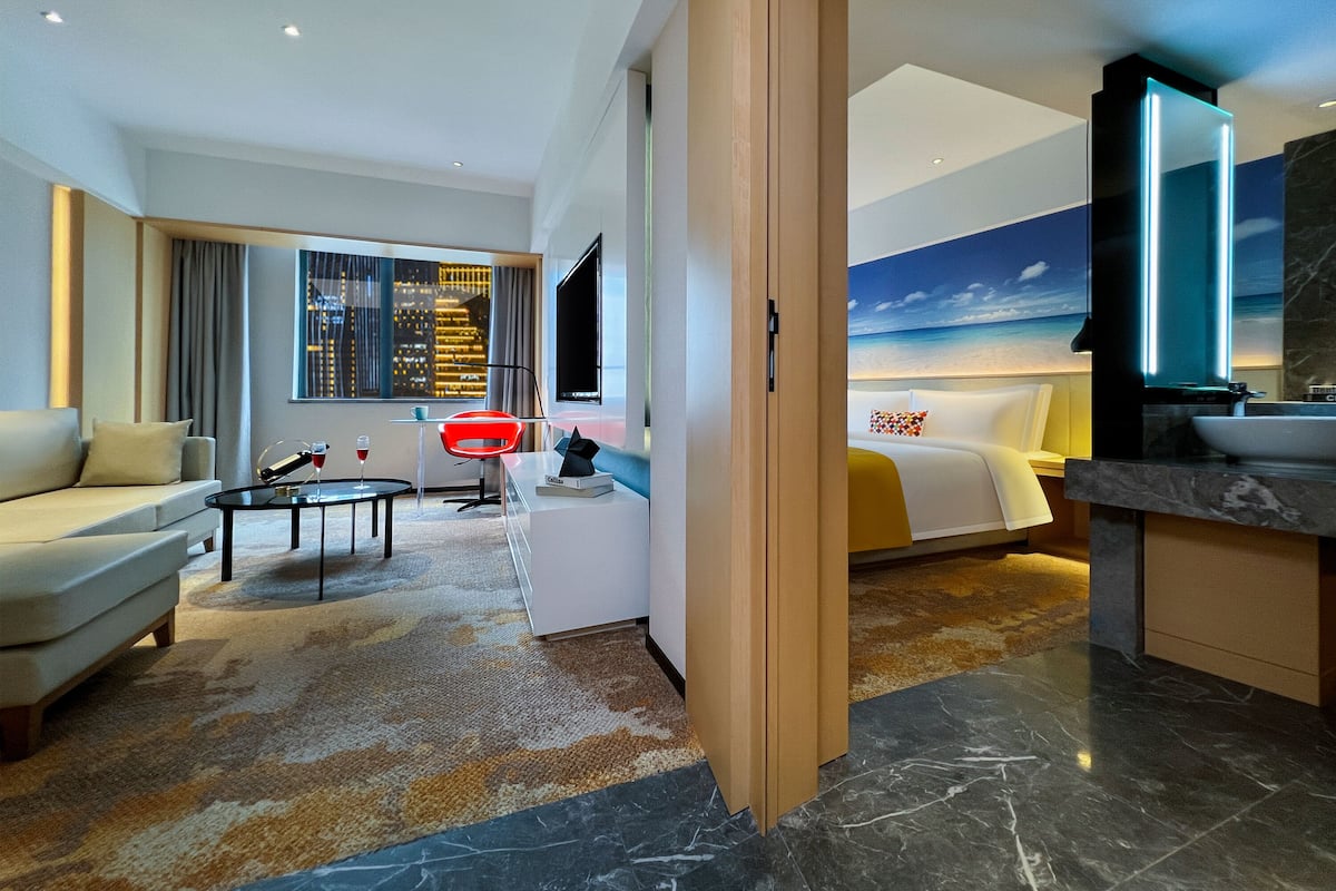 Ein modernes Hotelzimmer mit Sofa, Couchtisch, Fernseher, Bett und einem Badezimmer mit großem Spiegel.