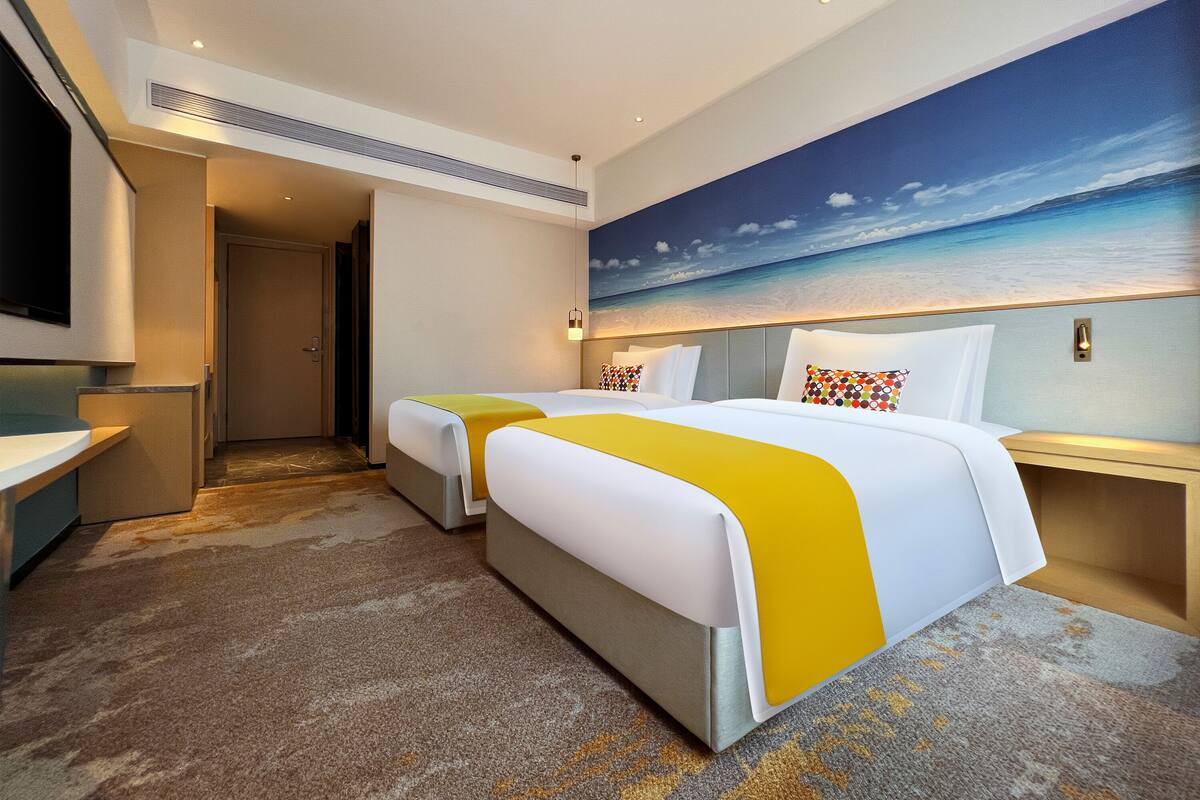 Ein Hotelzimmer mit zwei Betten, einem Strandgemälde, einem Fernseher und einem kleinen Schreibtisch.