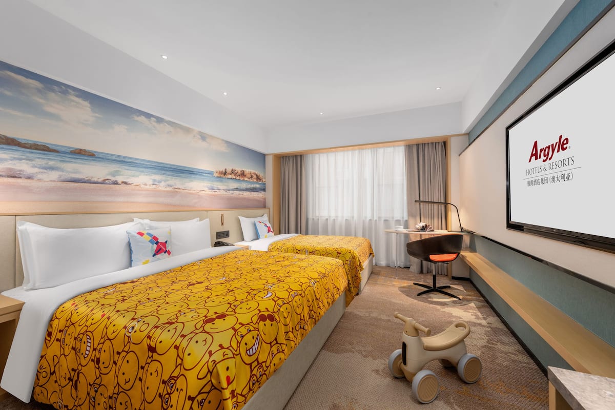 Ein Hotelzimmer mit zwei Betten, einem Schreibtisch, einem Stuhl, einer Projektionsleinwand und einem Wandbild mit einer Strandansicht.