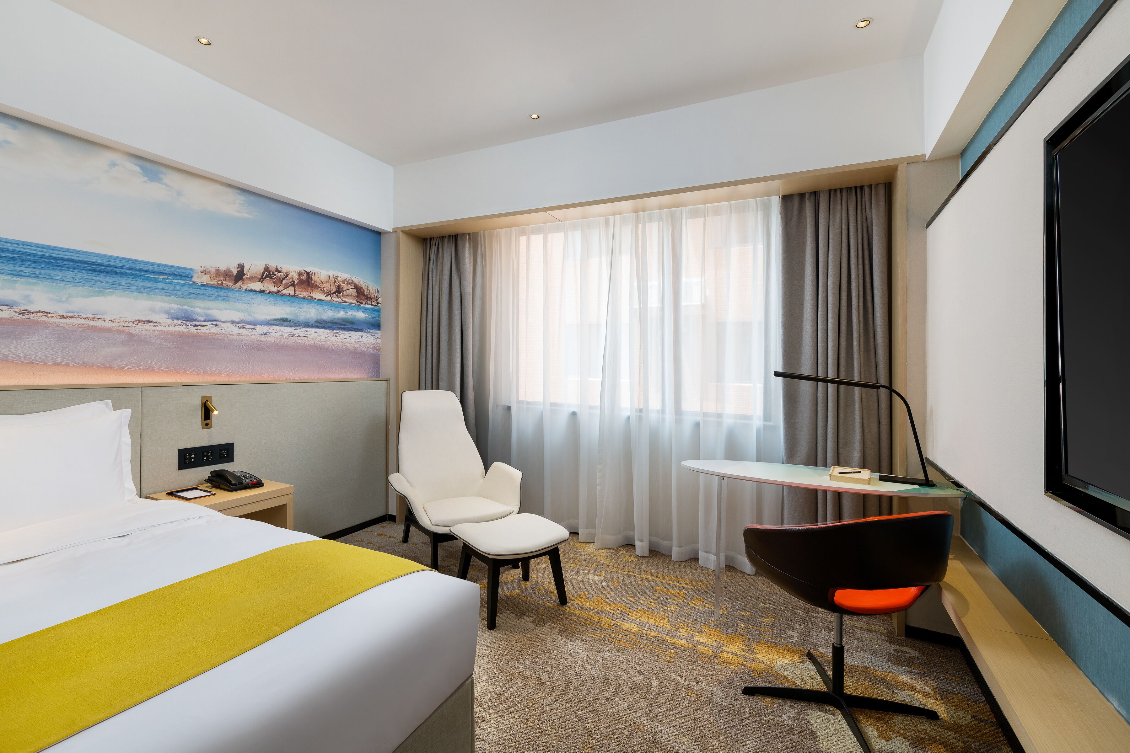 A hotel room with a bed, a desk, a chair, a TV, and a painting of a beach scene.