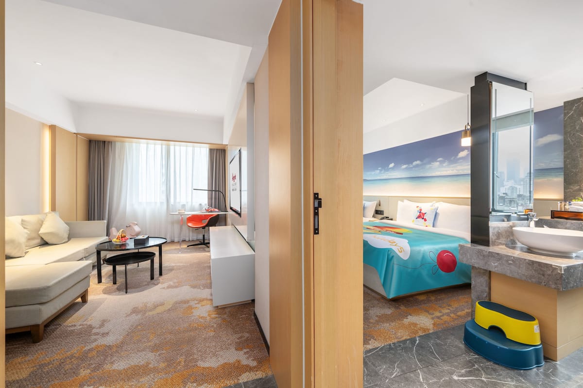 Ein Hotelzimmer mit Sofa, Esstisch, Bett mit farbenfroher Bettdecke, Badezimmer mit Waschbecken und Spiegel sowie einem Stadtbild-Wandgemälde.
