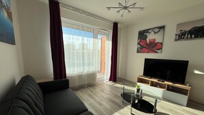 A modern living room with a black sofa, a TV on a wooden stand, a glass coffee table, and a large window with curtains. - Spacious 2BR + balcony in Prague I Comfort & Style. (Praha 9)