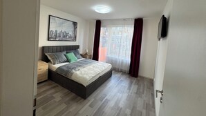 A bedroom with a bed, a nightstand, a lamp, a framed picture on the wall, and a window with curtains. - Spacious 2BR + balcony in Prague I Comfort & Style. (Praha 9)
