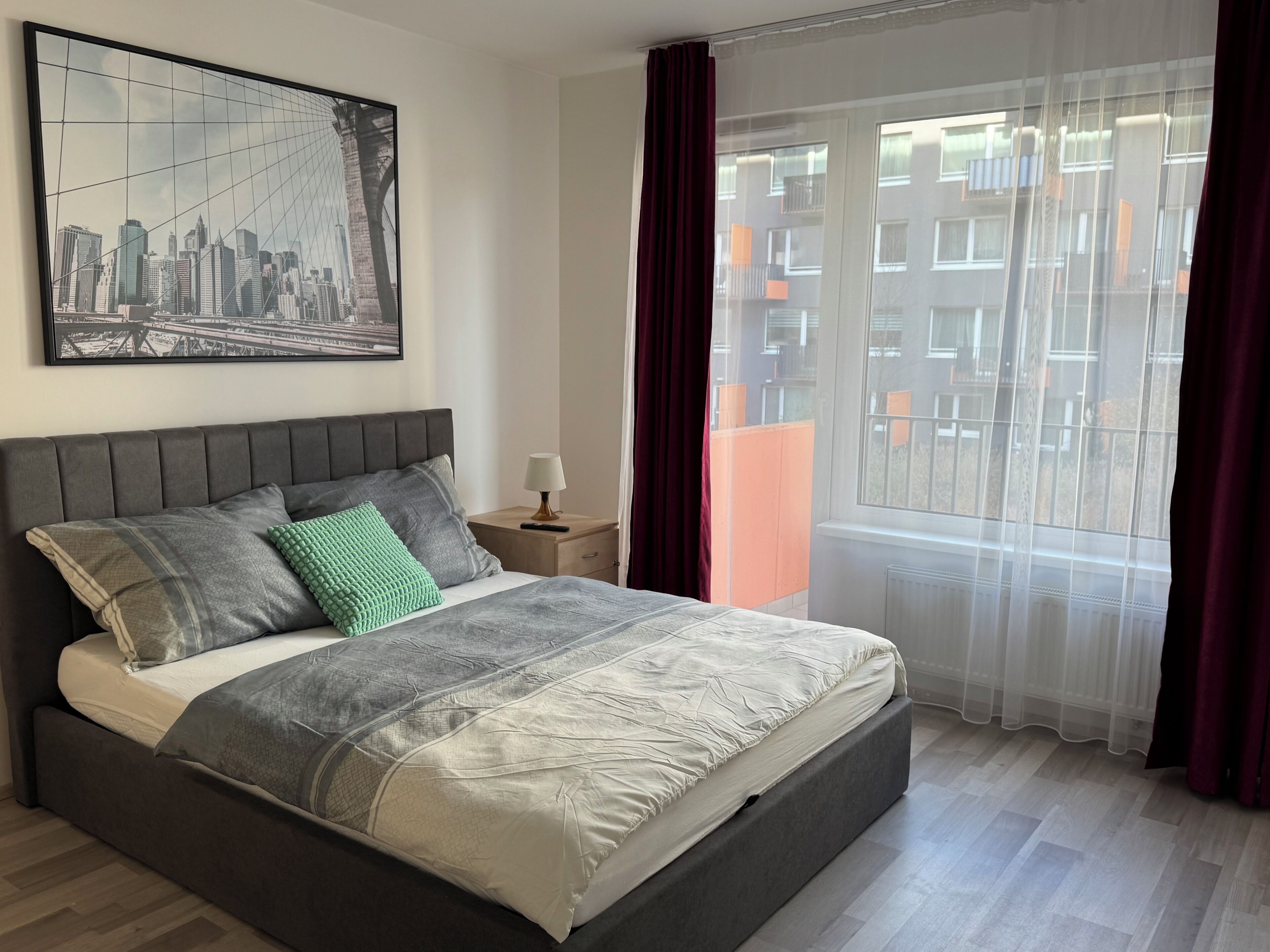 Een slaapkamer met een bed, een nachtkastje, een lamp, een raam met gordijnen en een stadslandschap op de muur.