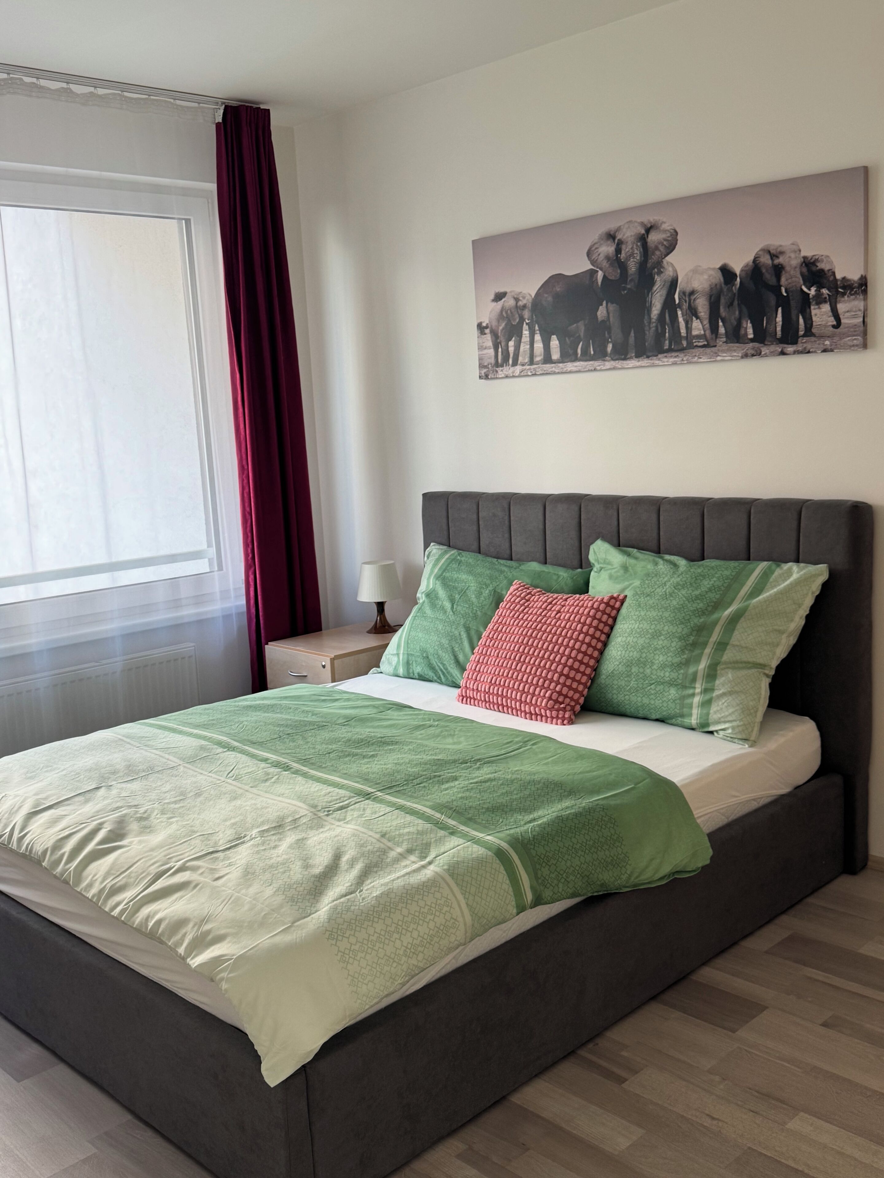 Ein Schlafzimmer mit einem Bett, grünen und roten Kissen, einem grauen Kopfteil, einem Fenster mit Vorhängen, einem Nachttisch mit einer Lampe und einem an der Wand hängenden Elefantenhänger.