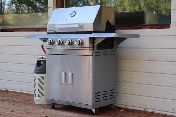 A portable stainless steel grill with four burners, a side compartment, and a chimney, placed on a wooden deck against a white building.
