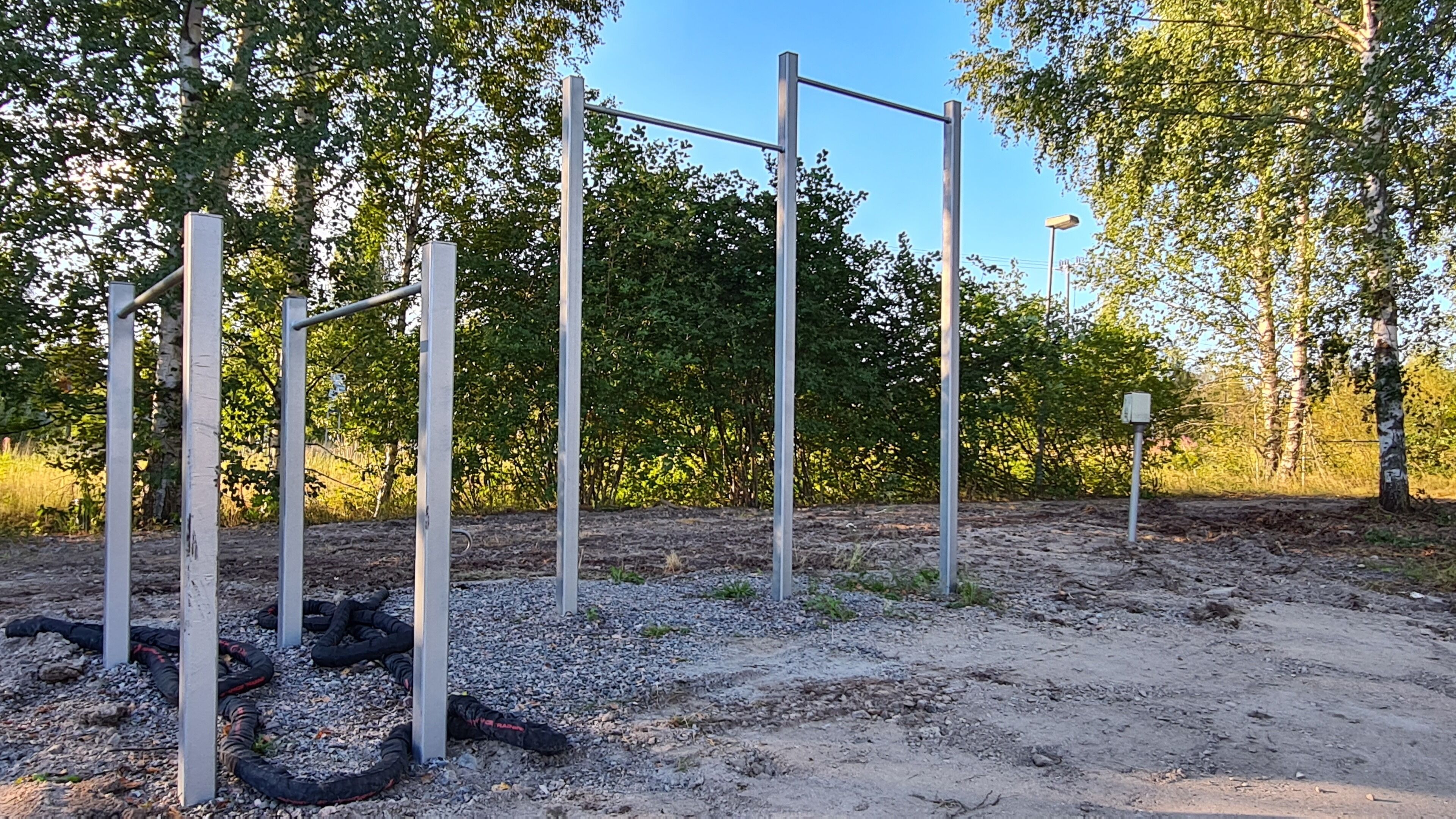 An outdoor gym with metal bars and ropes, surrounded by trees and gravel.