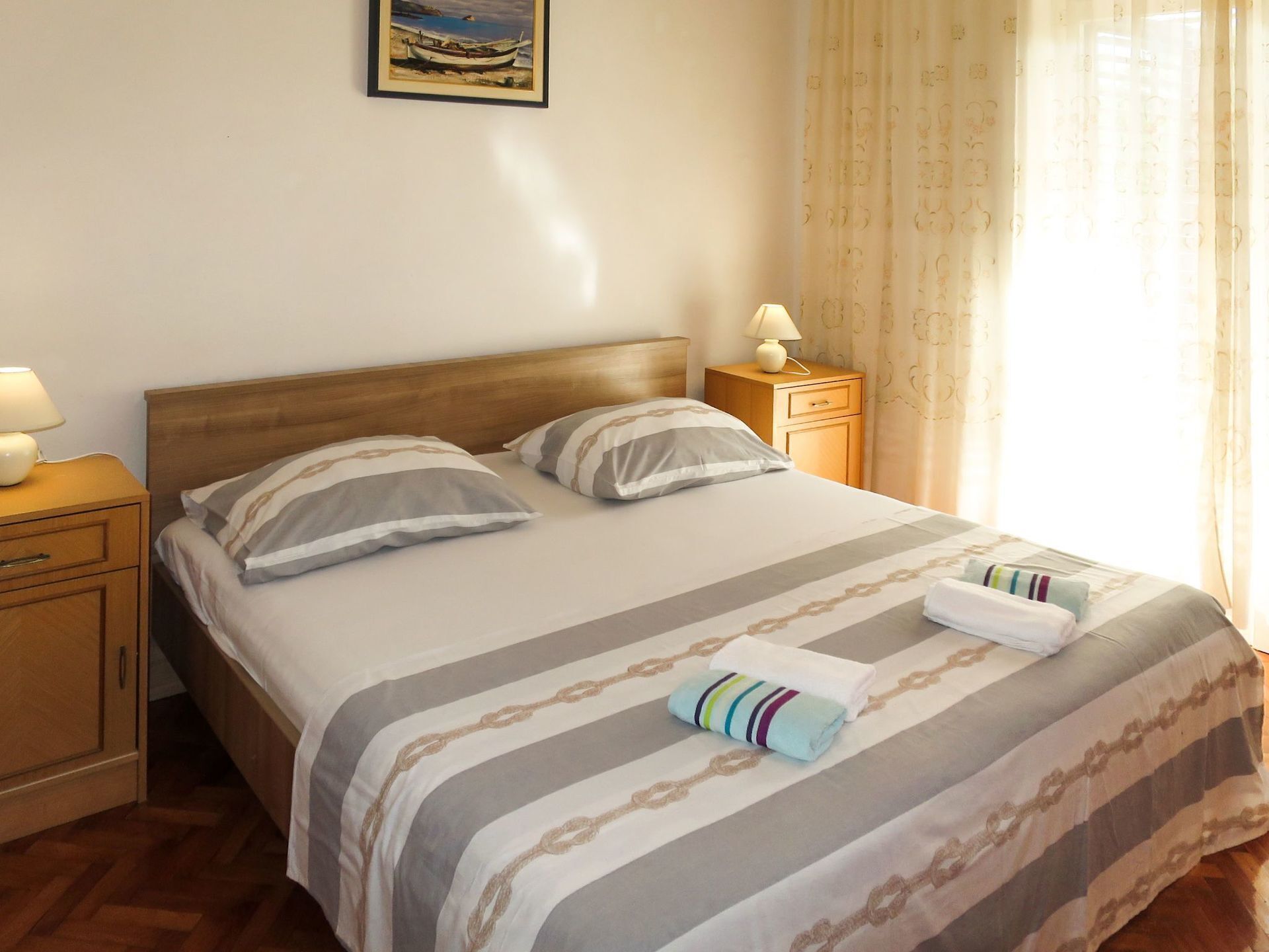 A neatly made bed with striped bedding, two pillows, a bedside table with a lamp, and a nightstand with a drawer.