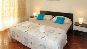 Ein ordentlich bezogenes Bett mit einer blumigen Tagesdecke, zwei blauen Kissen und einem Wandbild darüber.