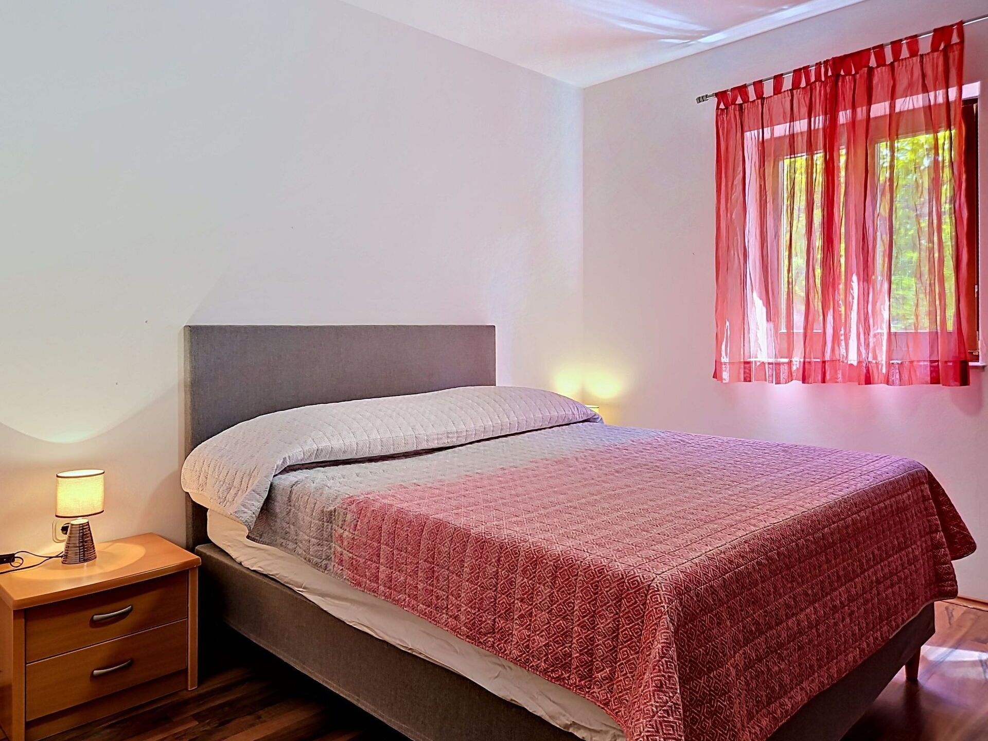 A neatly made bed with a patterned bedspread, a bedside lamp, and a window with sheer curtains.