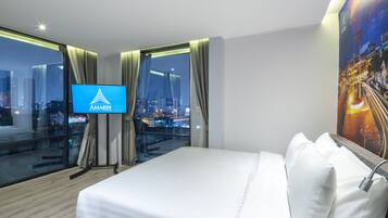 Hotelkamer met een groot bed, een televisie op een standaard, stadszicht door grote ramen en een schilderij aan de muur.