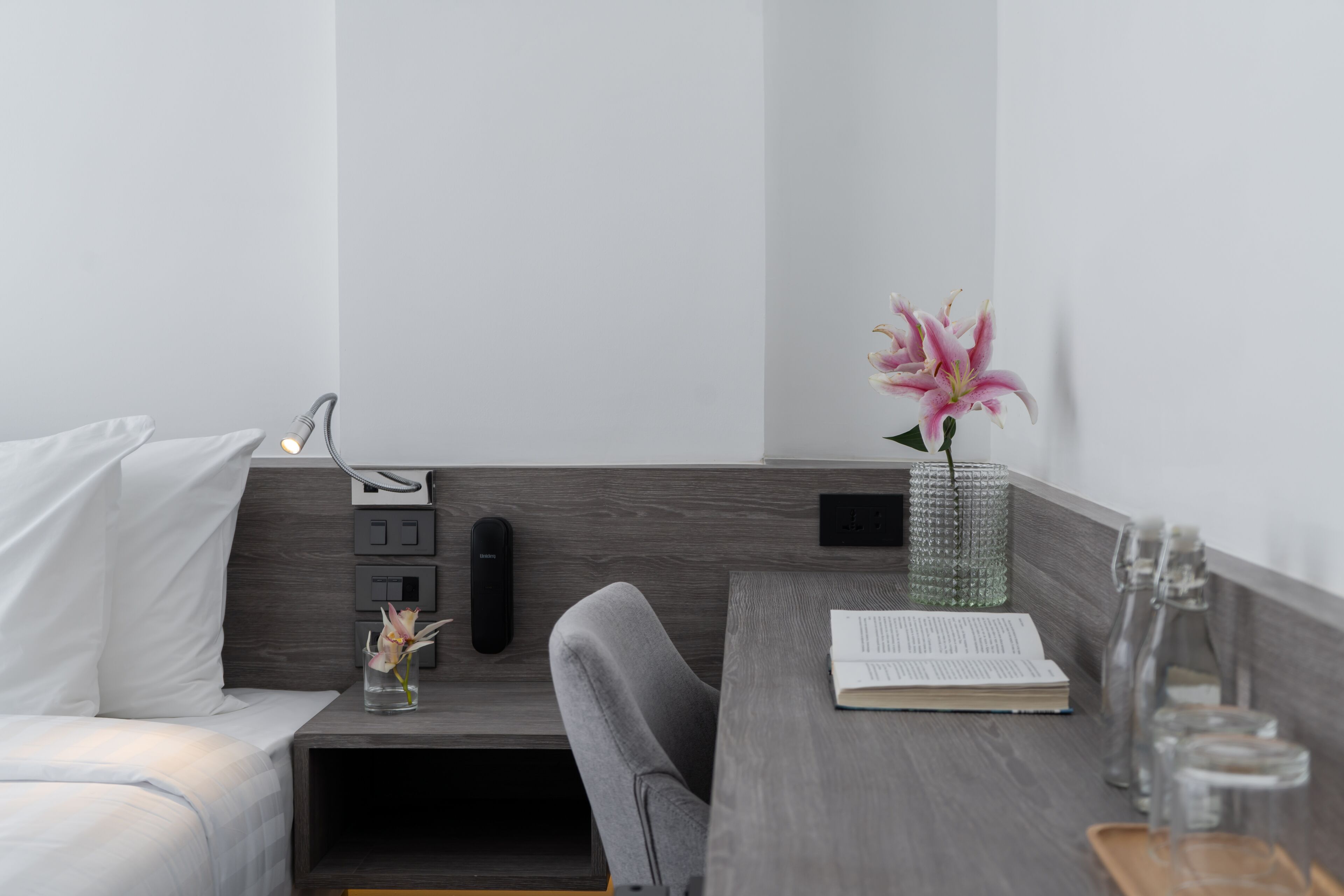 A modern bedroom with a bed, bedside table, and a desk area with a lamp, book, and vase with flowers.