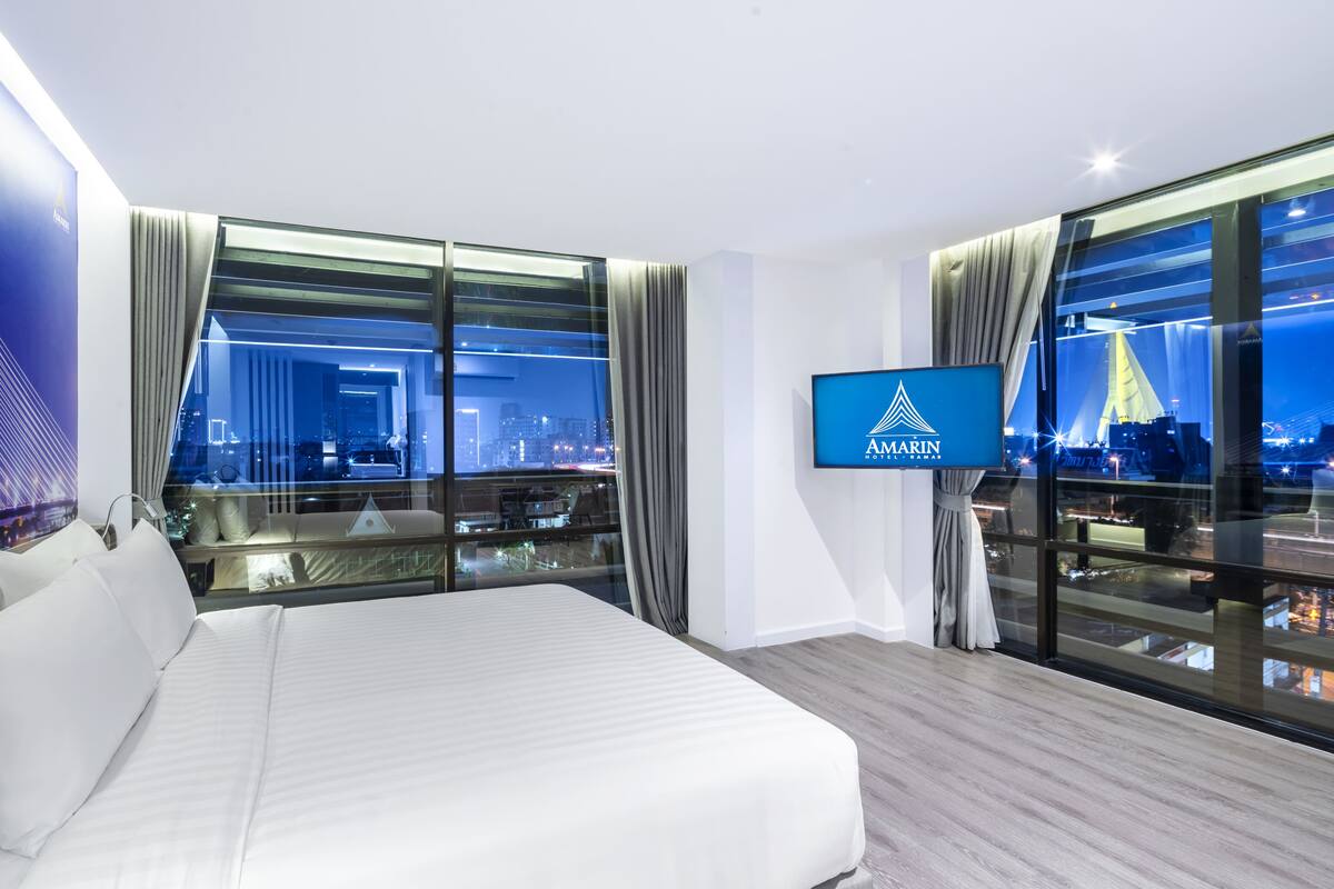A modern hotel room with a large bed, a TV, and a city view through floor-to-ceiling windows.