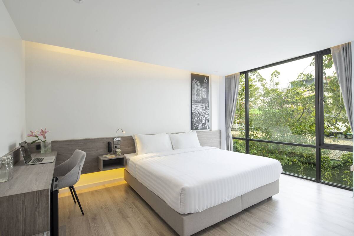 A modern hotel room with a large bed, a desk with a laptop, a chair, and a view of greenery through large windows.