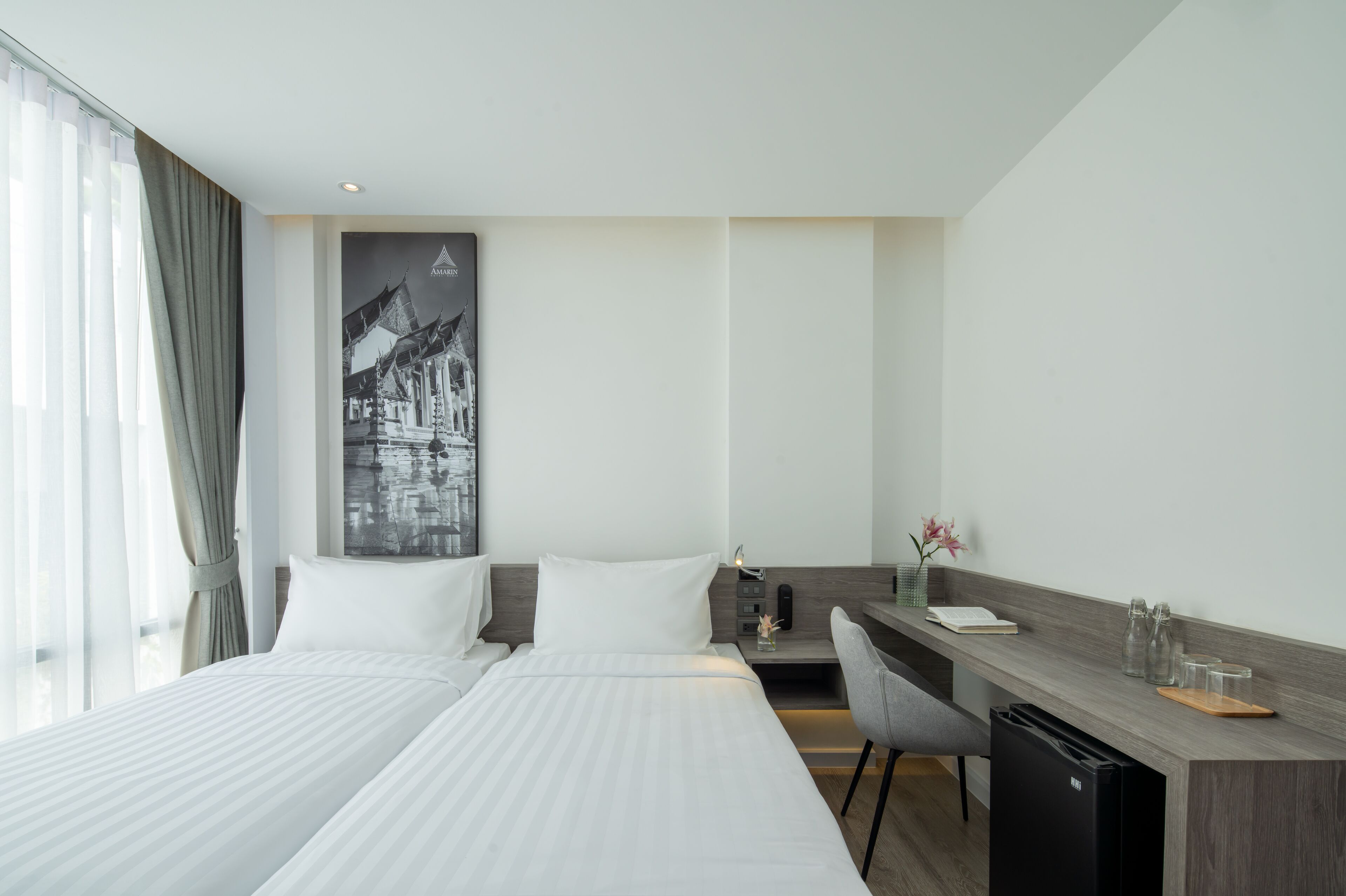 A hotel room with a bed, a desk, a chair, a small fridge, and a large window with curtains.