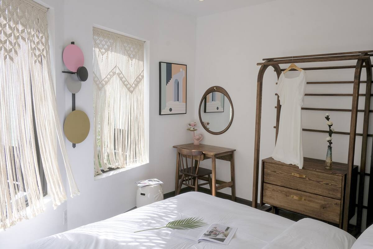 A bedroom with a bed, a wooden dresser, a chair, a small table, a mirror, and a window with curtains.
