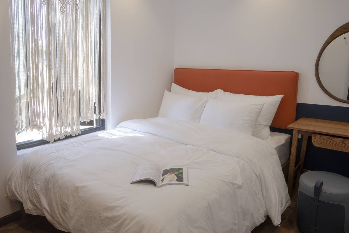 A neatly made bed with white bedding and pillows, a wooden nightstand with a mirror, and a window with sheer curtains.