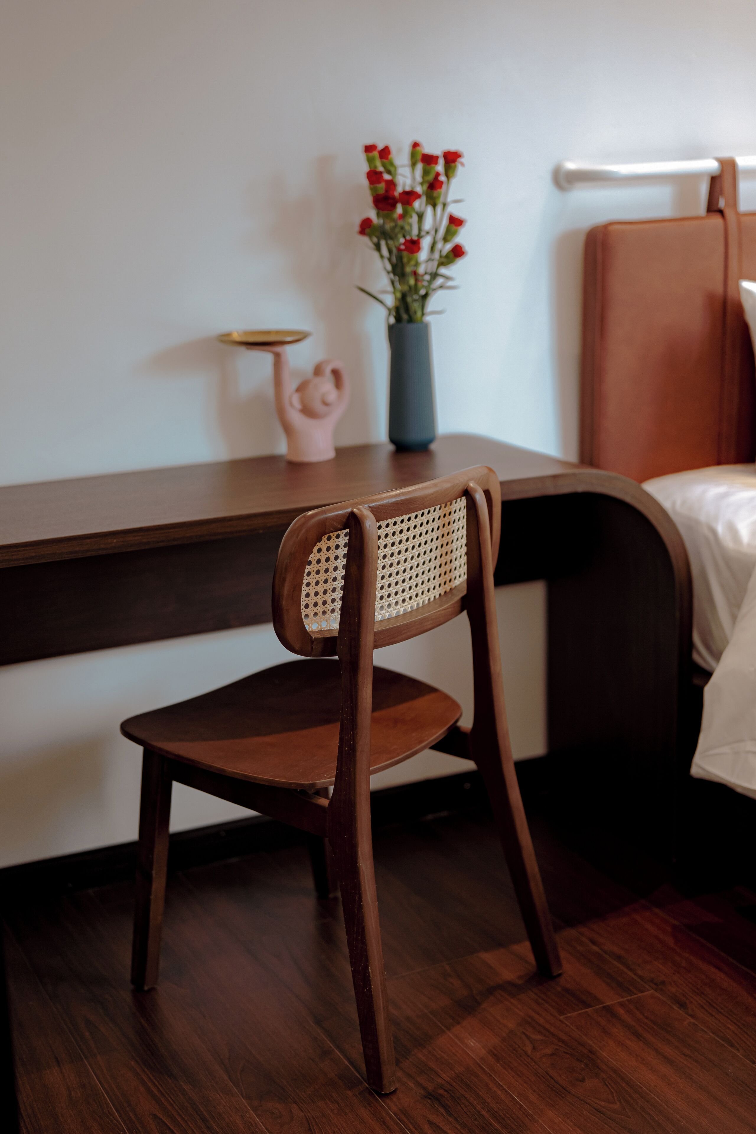 Una silla de madera con respaldo tejido, una mesita con un jarrón de flores rojas y una cama con cabecera marrón.
