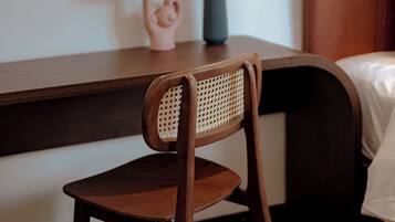 Una silla de madera con respaldo tejido, una mesita con un jarrón de flores rojas y una cama con cabecera marrón.