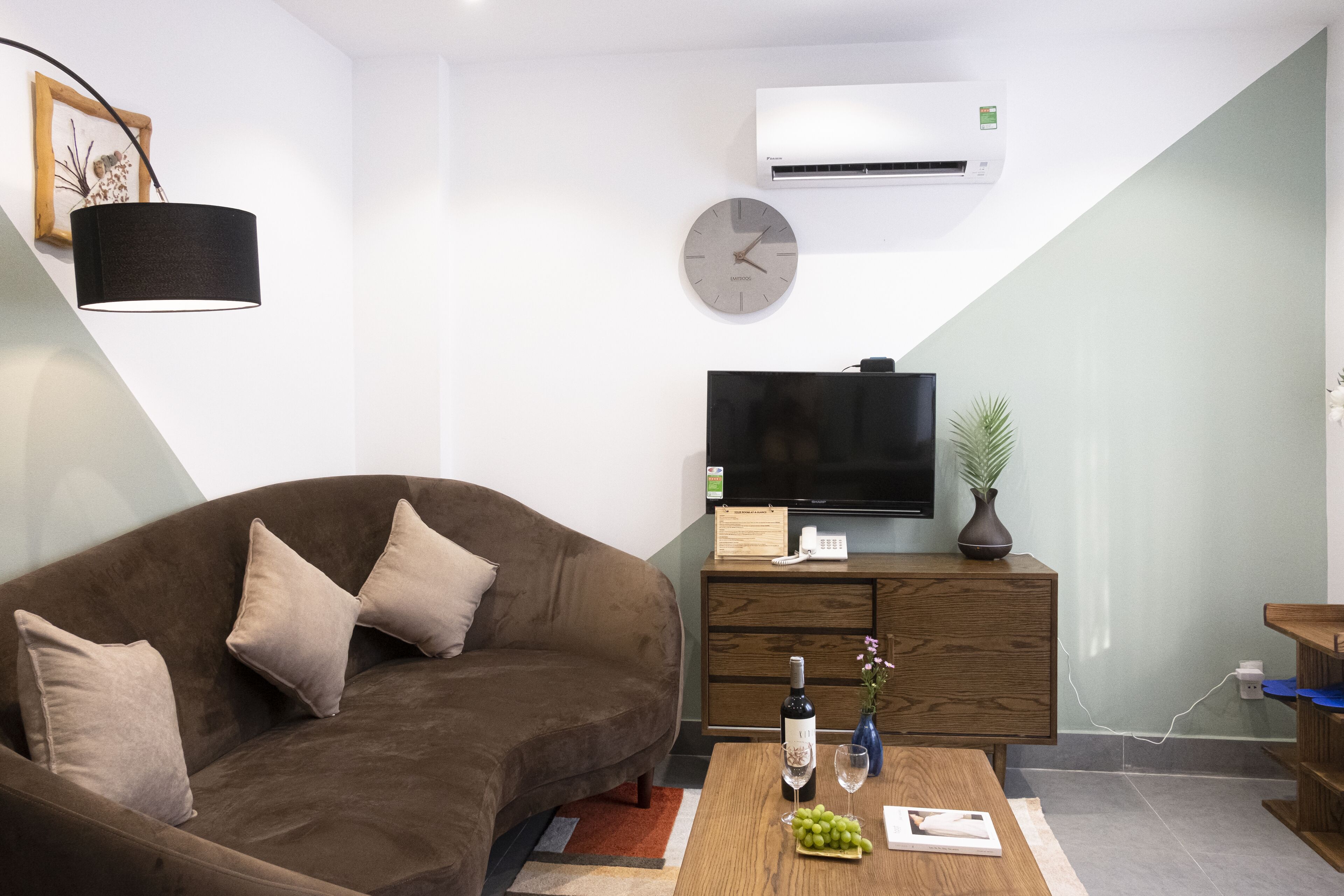 A modern living room with a brown sofa, a flat-screen TV, a wooden cabinet, and a coffee table with a bottle of wine and a vase of flowers.