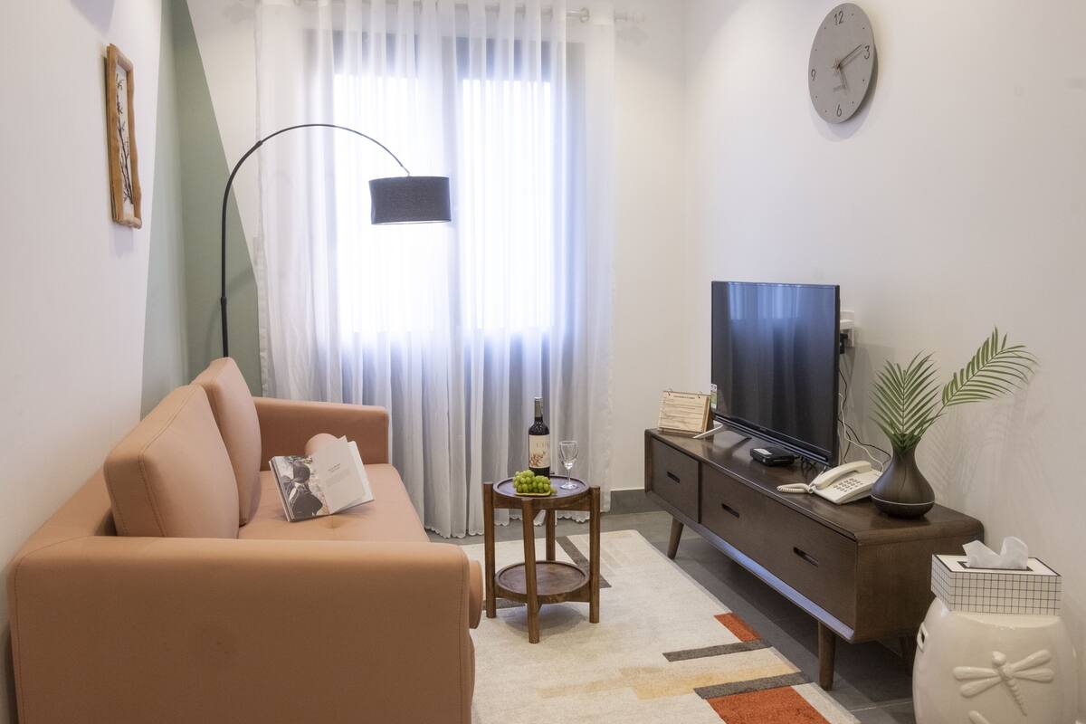 A living room with a sofa, a television, a small table with a bottle and glasses, a clock, and a plant.
