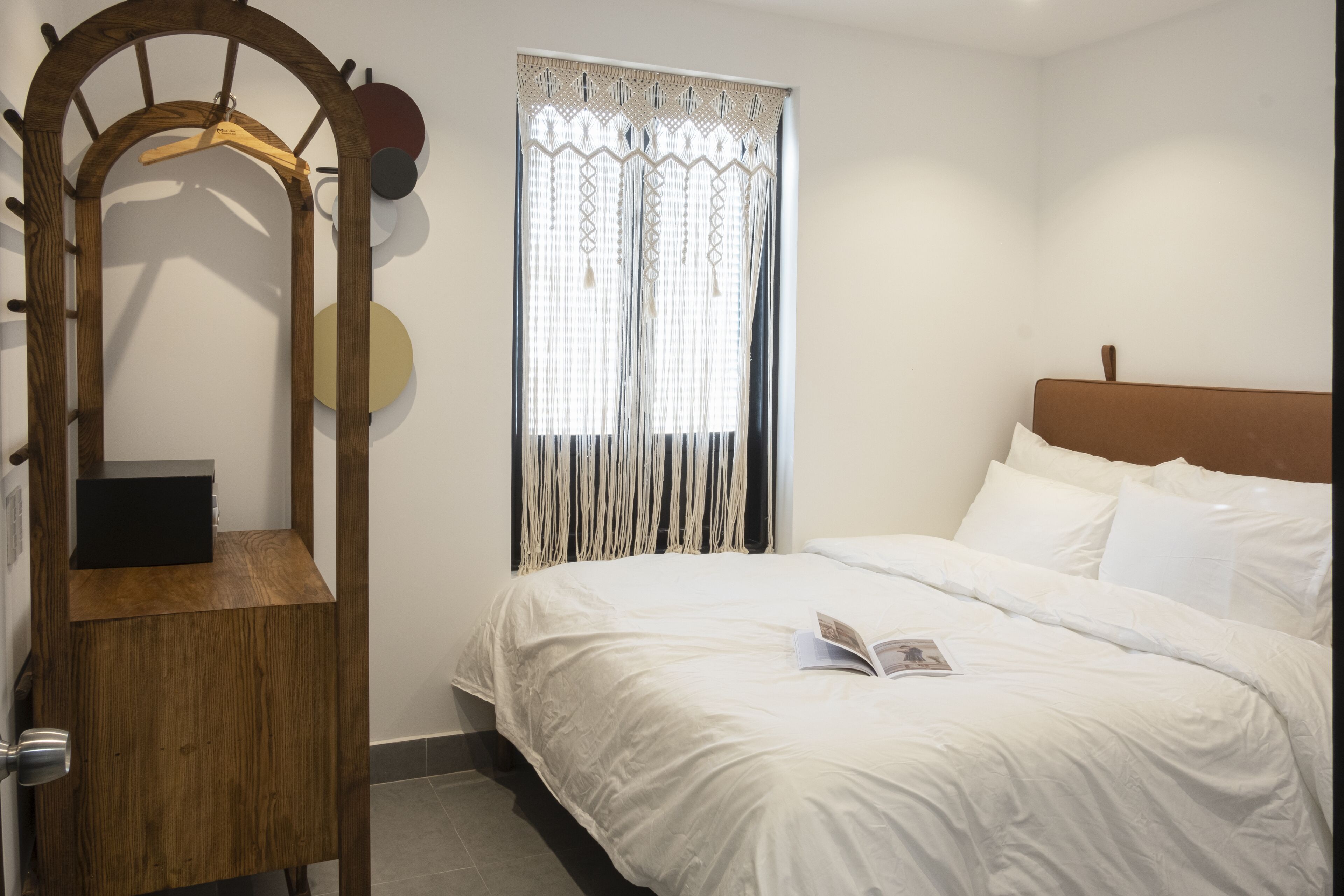 Un dormitorio con un armario de madera, una cama con ropa de cama blanca y una ventana con cortinas estampadas.