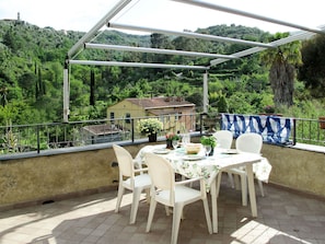 A covered outdoor patio with a table set for two, surrounded by white chairs, overlooking a lush green landscape with a building in the distance.
