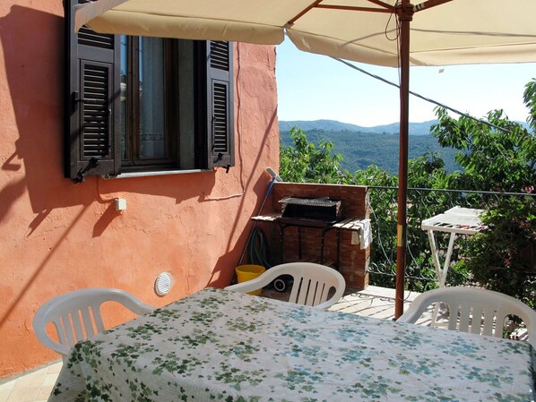 A patio with a table covered by a floral tablecloth, two white plastic chairs, a barbecue grill, and a view of the mountains. - Beautiful private villa for 5 guests with pool, TV, terrace and pets allowed (Dolcedo)