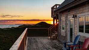 A wooden deck with a view of the ocean at sunset, a covered area with Adirondack chairs, and a building with a wooden exterior.
