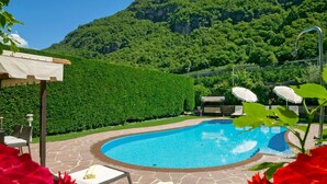 A swimming pool with lounge chairs and umbrellas, surrounded by a hedge and a mountainous backdrop.