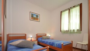 A room with two wooden beds, a small table, a chair, a window with curtains, and a framed picture on the wall. - Beautiful  villa for 6 people with A/C, TV, terrace and pets allowed (Lignano Sabbiadoro)